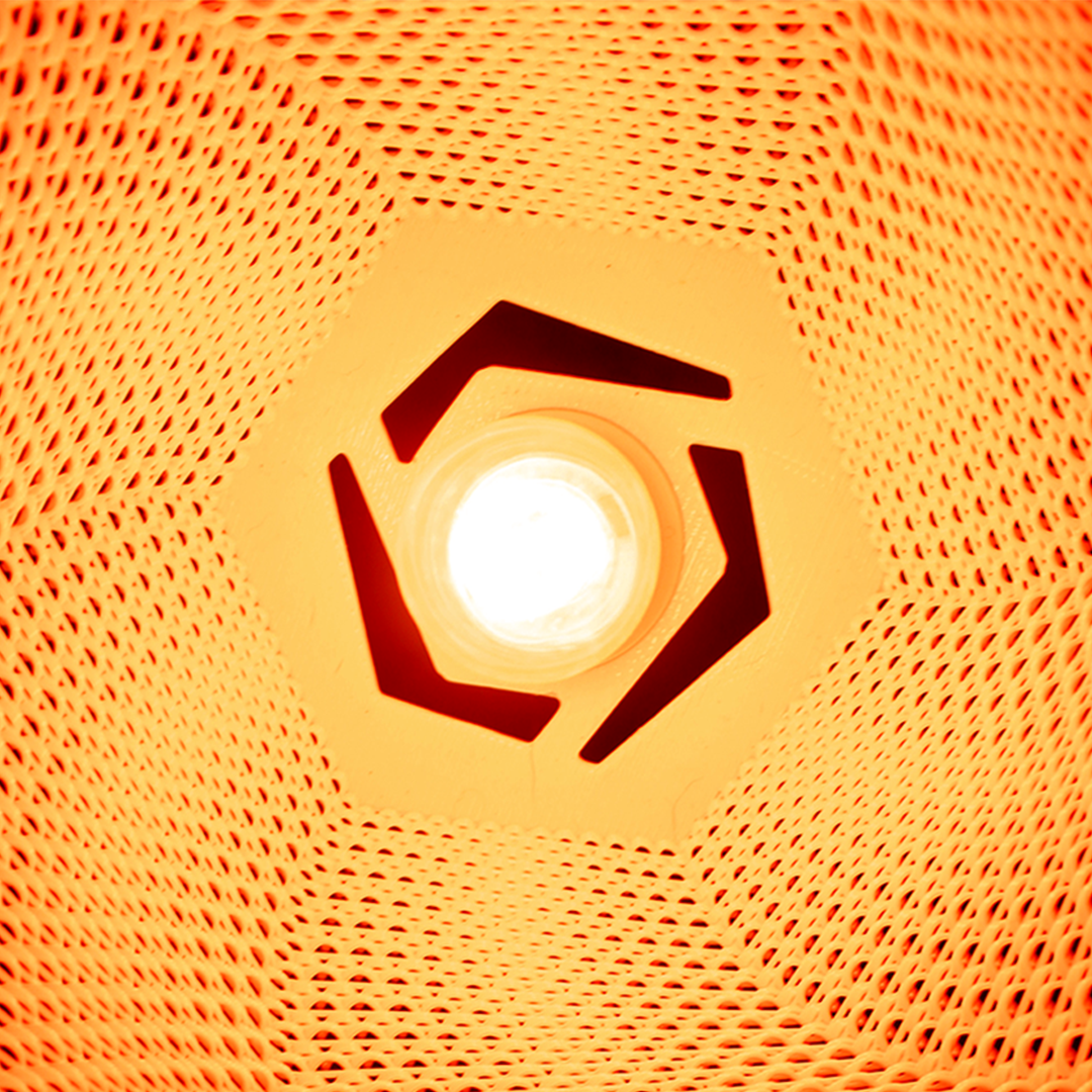 A glowing light bulb is seen from inside an orange mesh lampshade with a hexagonal opening and geometric cutouts
