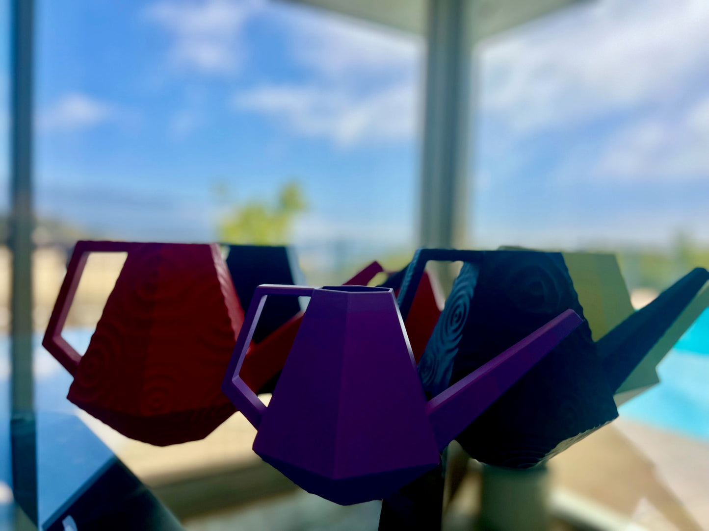 Geometric watering cans in vibrant colors, showcased against a blurred outdoor backdrop with a pool and sky view.