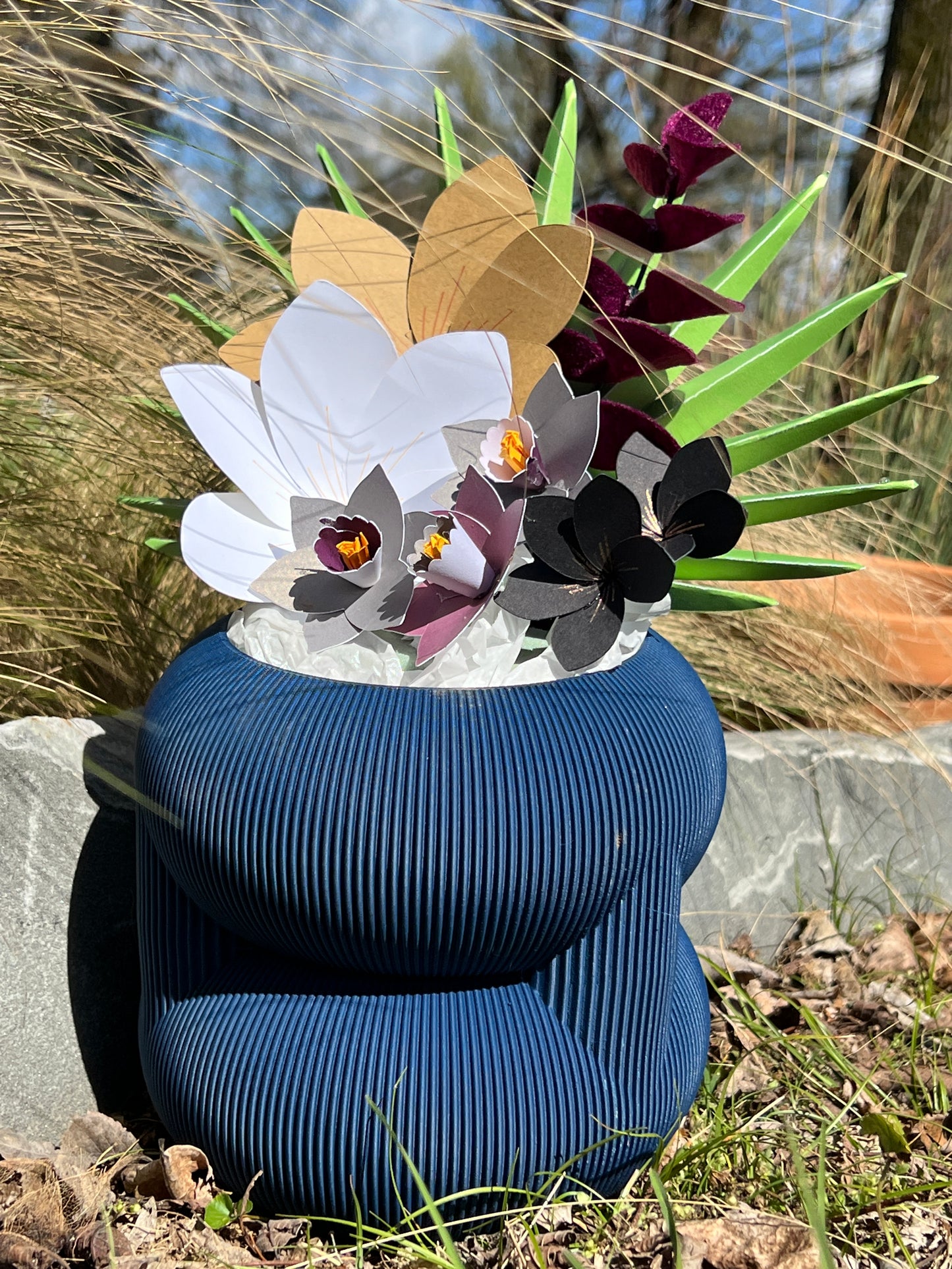 A blue rounded stacked planter filled with decorative paper flowers, positioned outdoors to create a charming natural aesthetic.