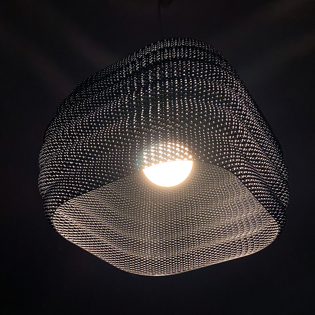 Daira Lampshade beautifully illuminated, surrounded by a dark backdrop, highlighting its intricate woven texture.