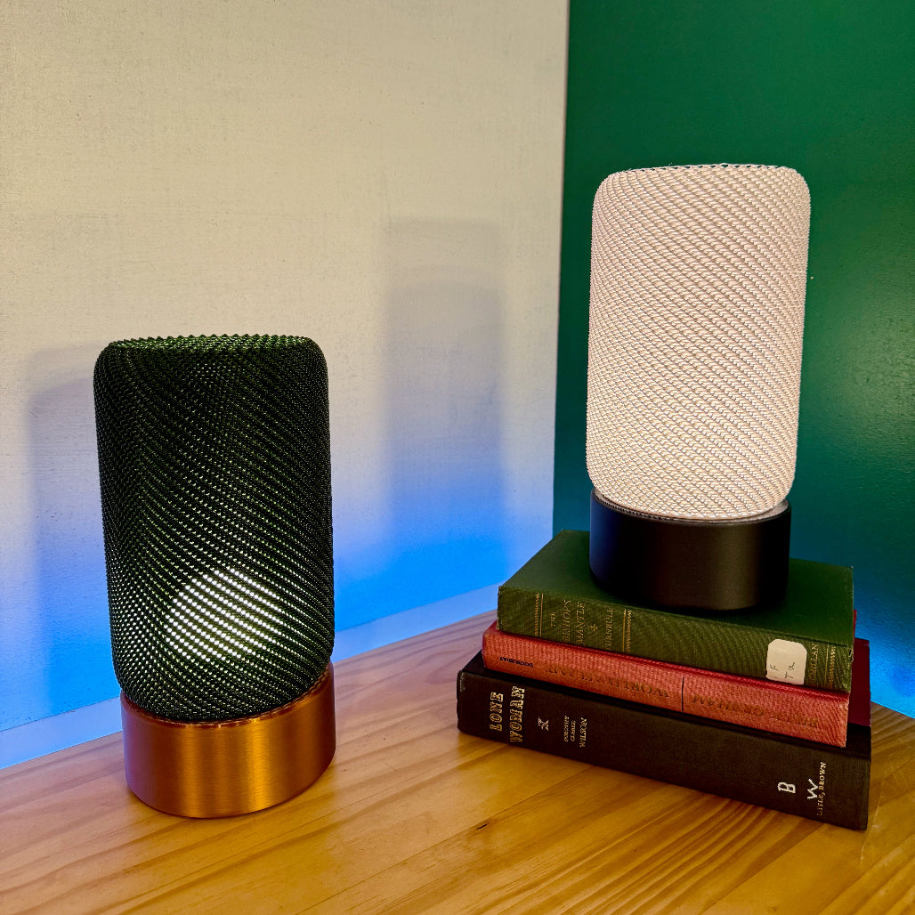 Two EVE Table Lamps, one green and one white, casting warm light while resting on stacked books with an ambient blue glow in the background.