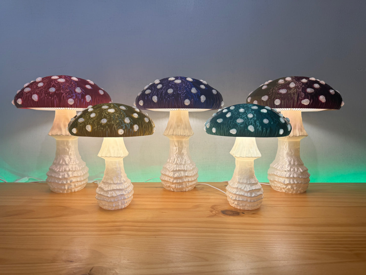 A collection of five vibrant Amanita fungi lamps in various colors, showcasing their whimsical design with glowing caps and intricate details on a wooden table.