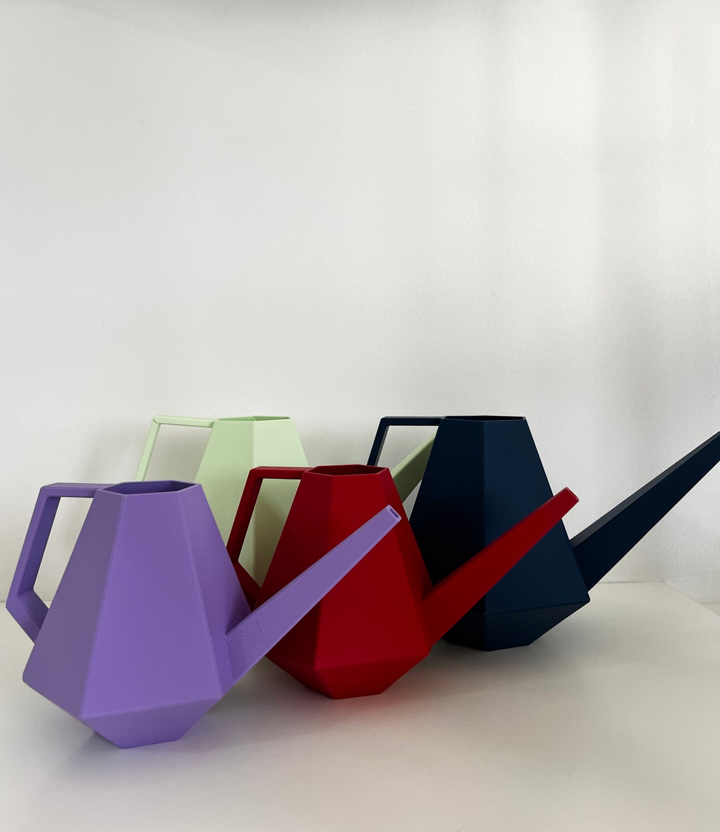 Colorful geometric watering cans in various matte finishes, including purple, green, red, and blue, arranged on a white surface.