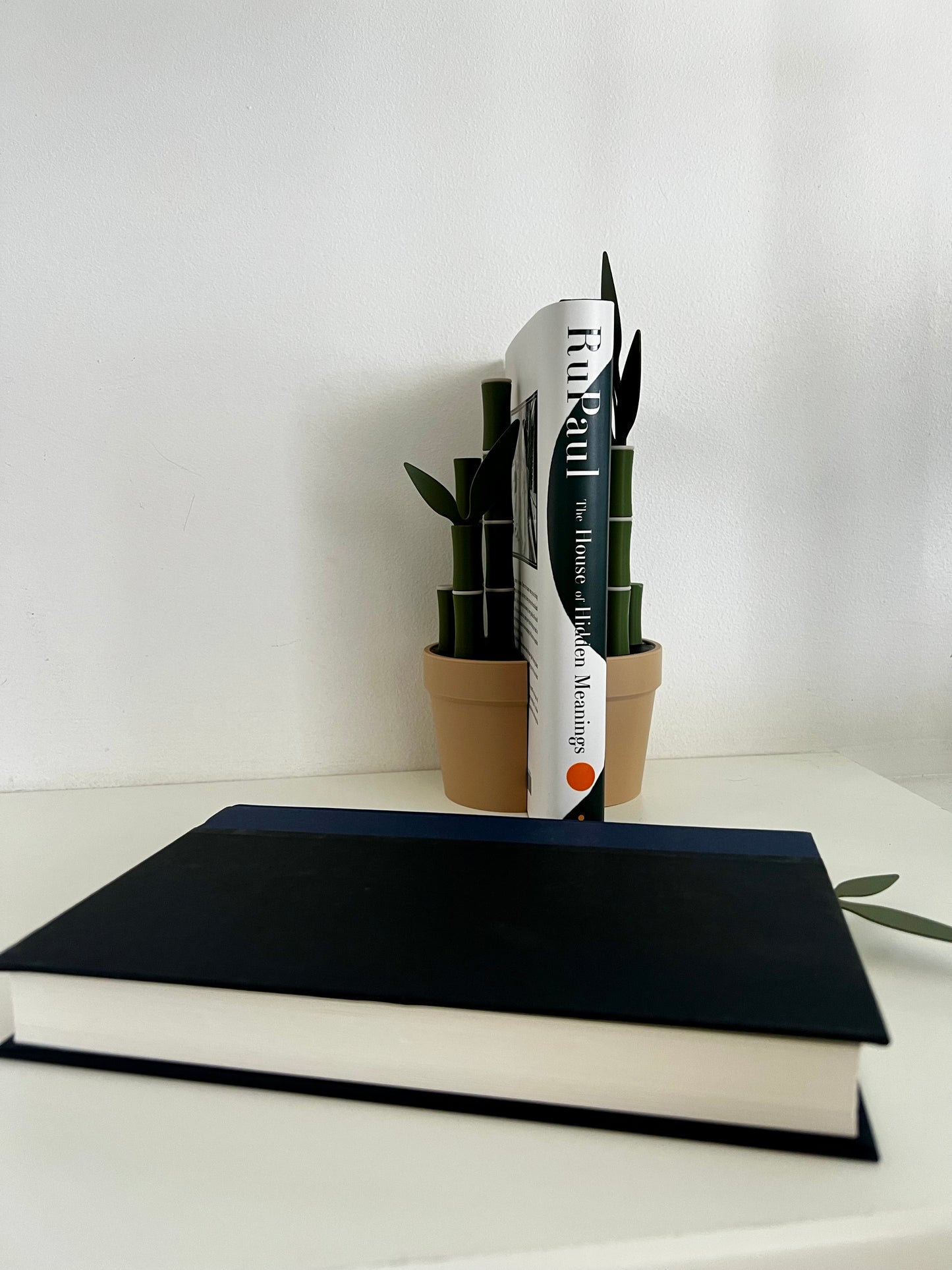 Side view of a bamboo bookend supporting the RuPaul book with a solid blue book below, all arranged on a minimalist white table.
