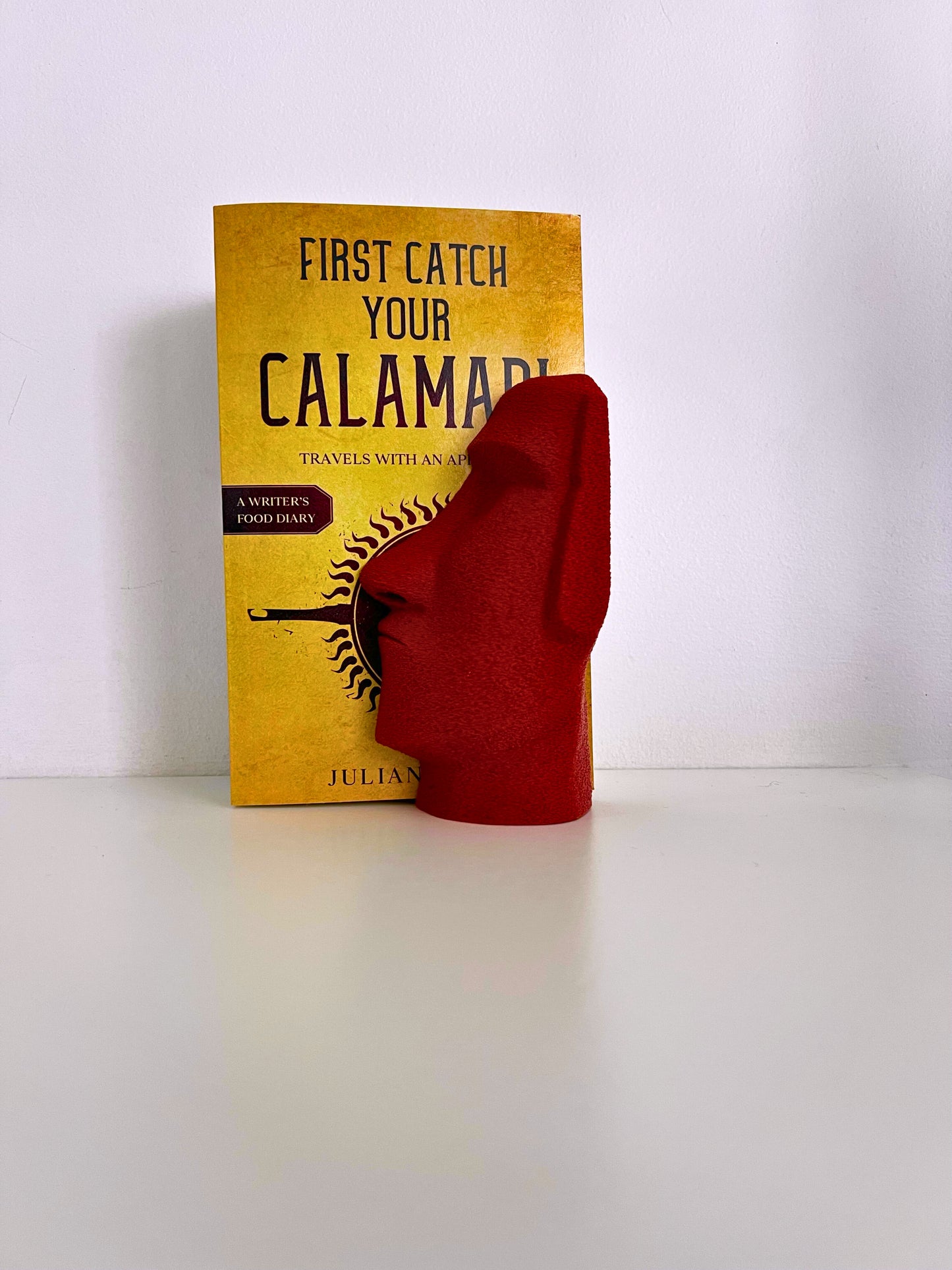 Red sculptural Moai bookend displaying alongside a cookbook, merging functionality and artistic flair.