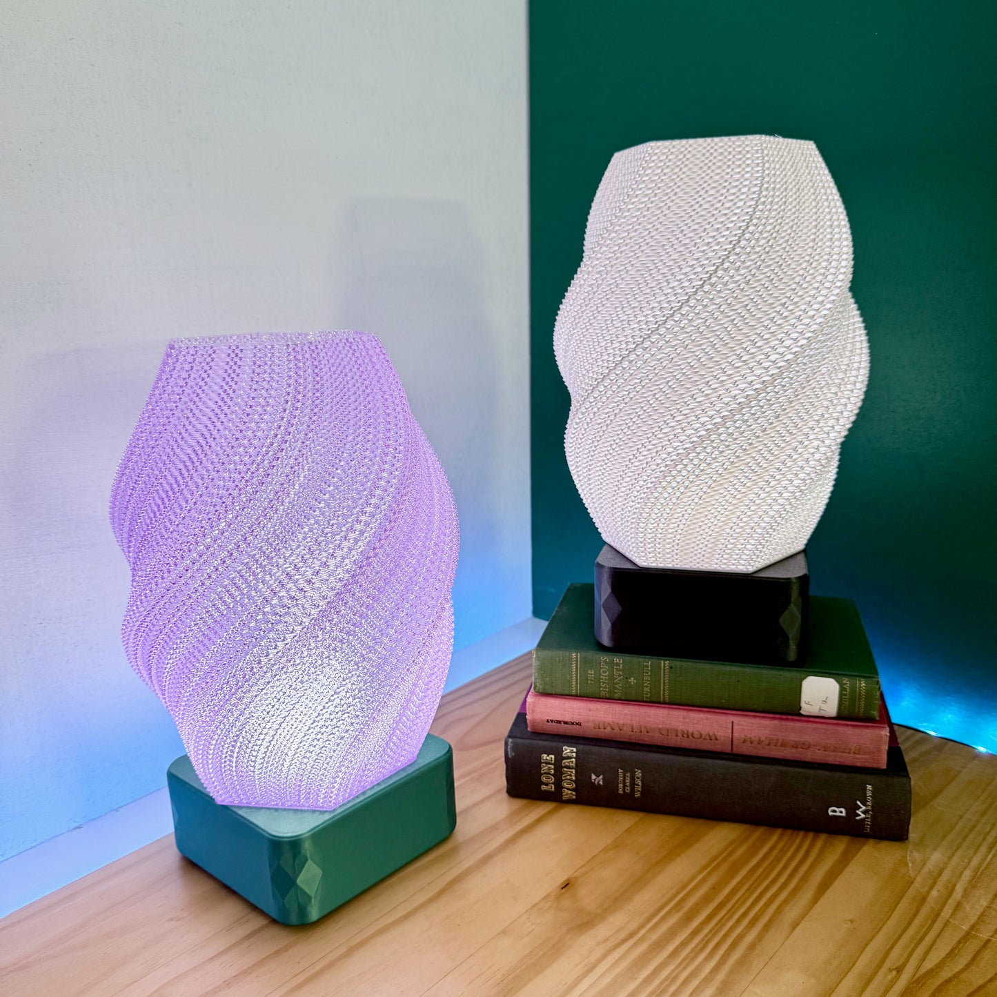 A white and a purple spiral-textured lamp sit on a wooden surface, with books stacked under the white lamp.