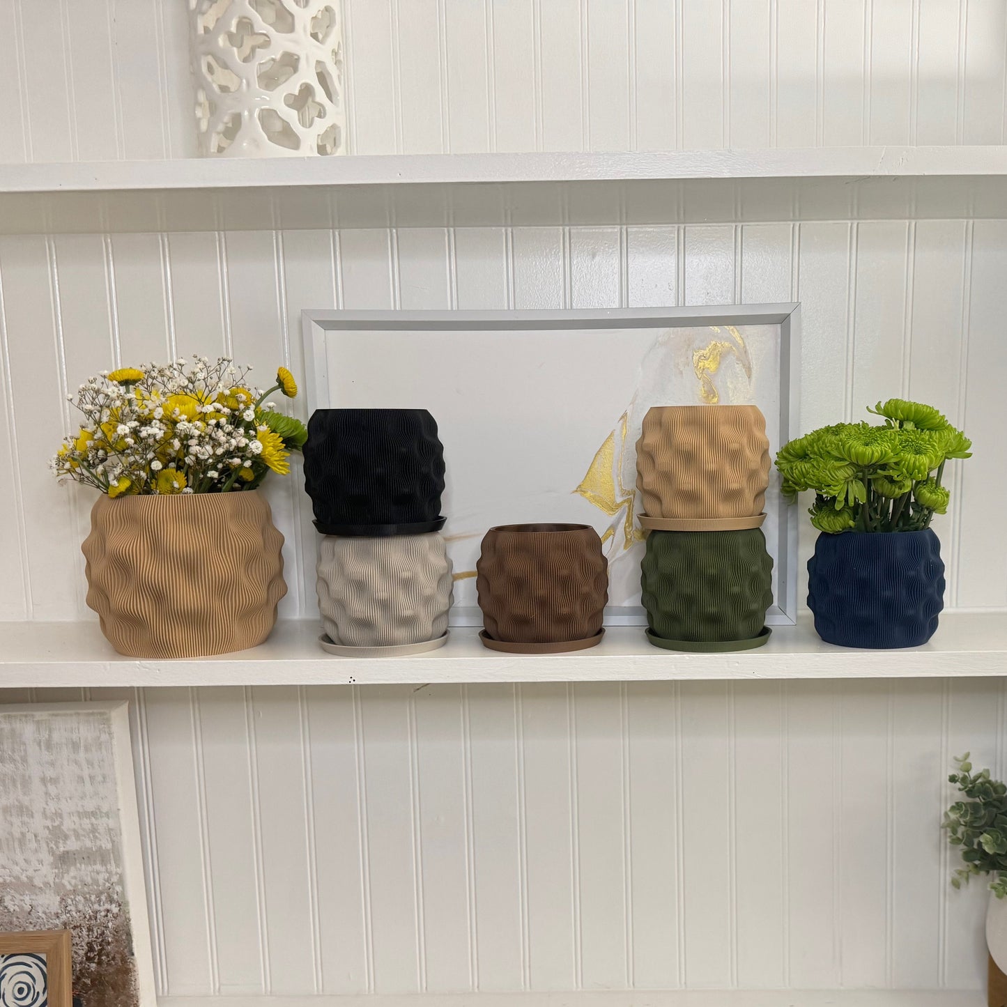 A collection of matte dimpled planters displayed on a shelf, showcasing different sizes and colors with beautiful flowers.