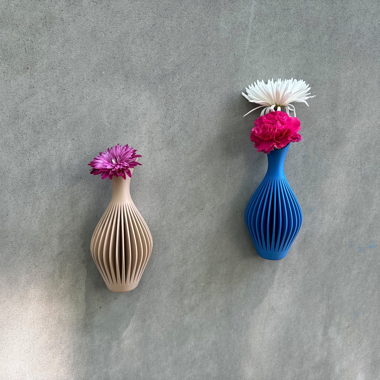 Two unique hanging wall vases with modern ribbed design, one beige and one blue, each featuring vibrant flowers, displayed on a textured gray wall.