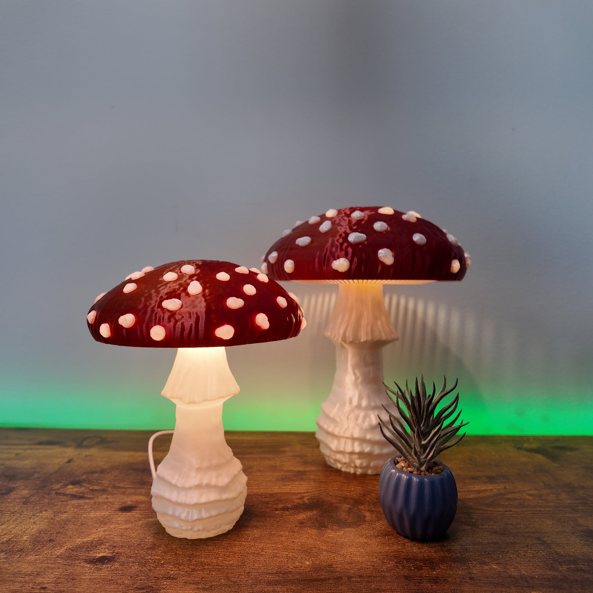 Two shimmering Amanita fungi lamps with red caps and white spots, illuminating a wooden surface with a soft glow, surrounded by a decorative plant.