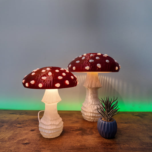 Two shimmering Amanita fungi lamps with red caps and white spots, illuminating a wooden surface with a soft glow, surrounded by a decorative plant.