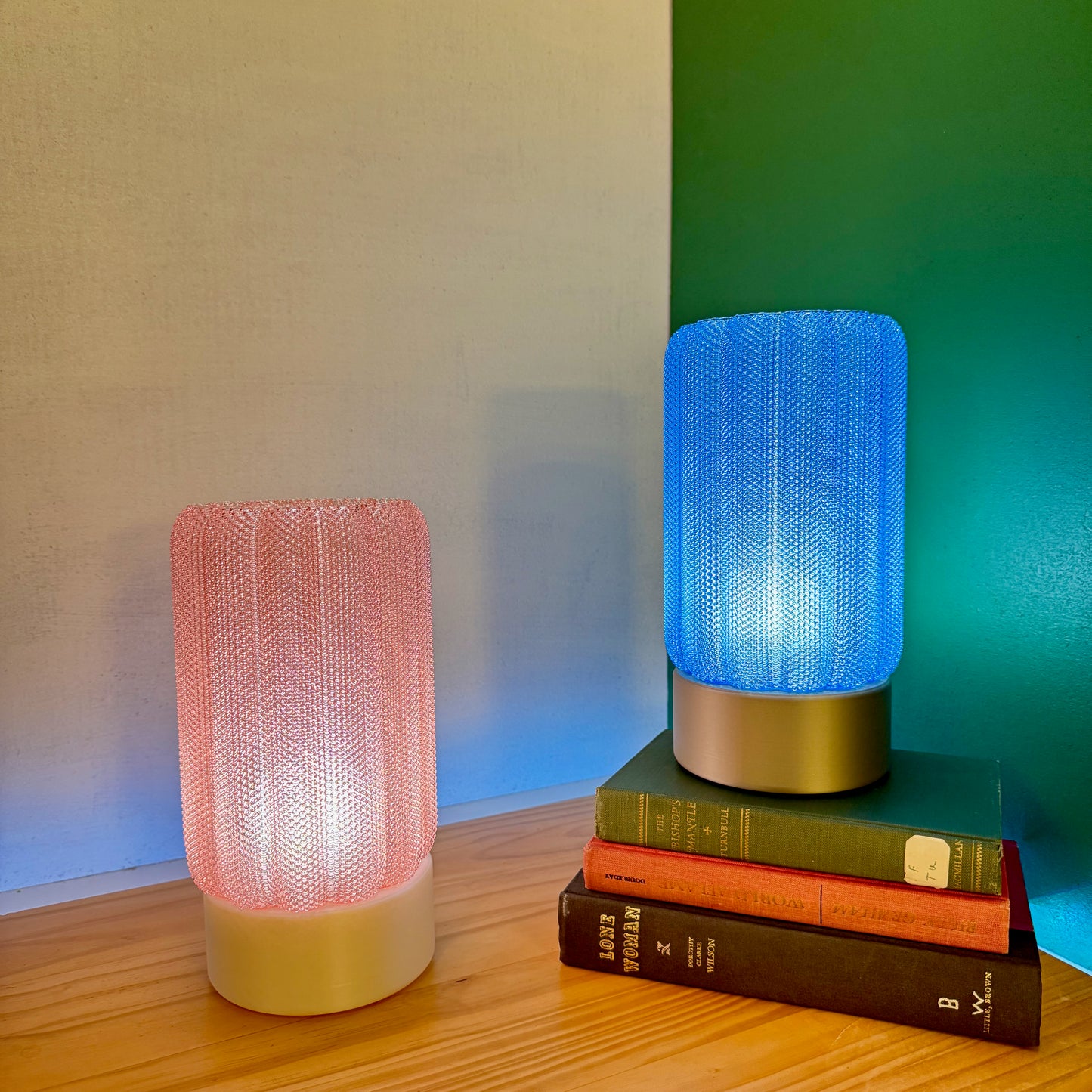 A pink and a blue mesh-covered lamp glow softly on a wooden surface, with books stacked under the blue lamp.