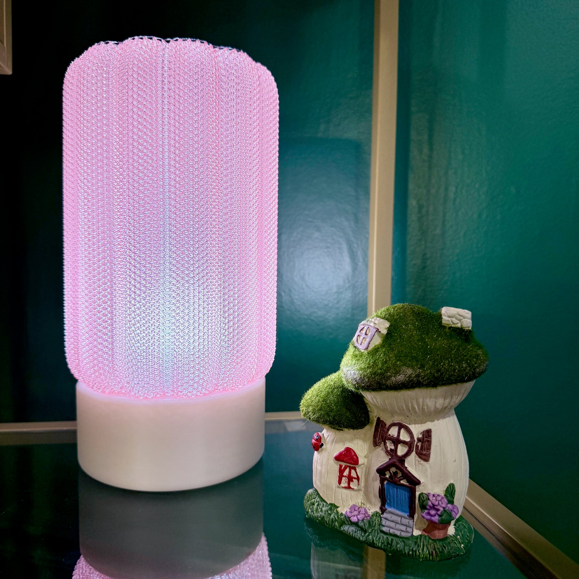 A pink mesh lamp sits next to a whimsical mushroom-shaped house ornament on a glass shelf.
