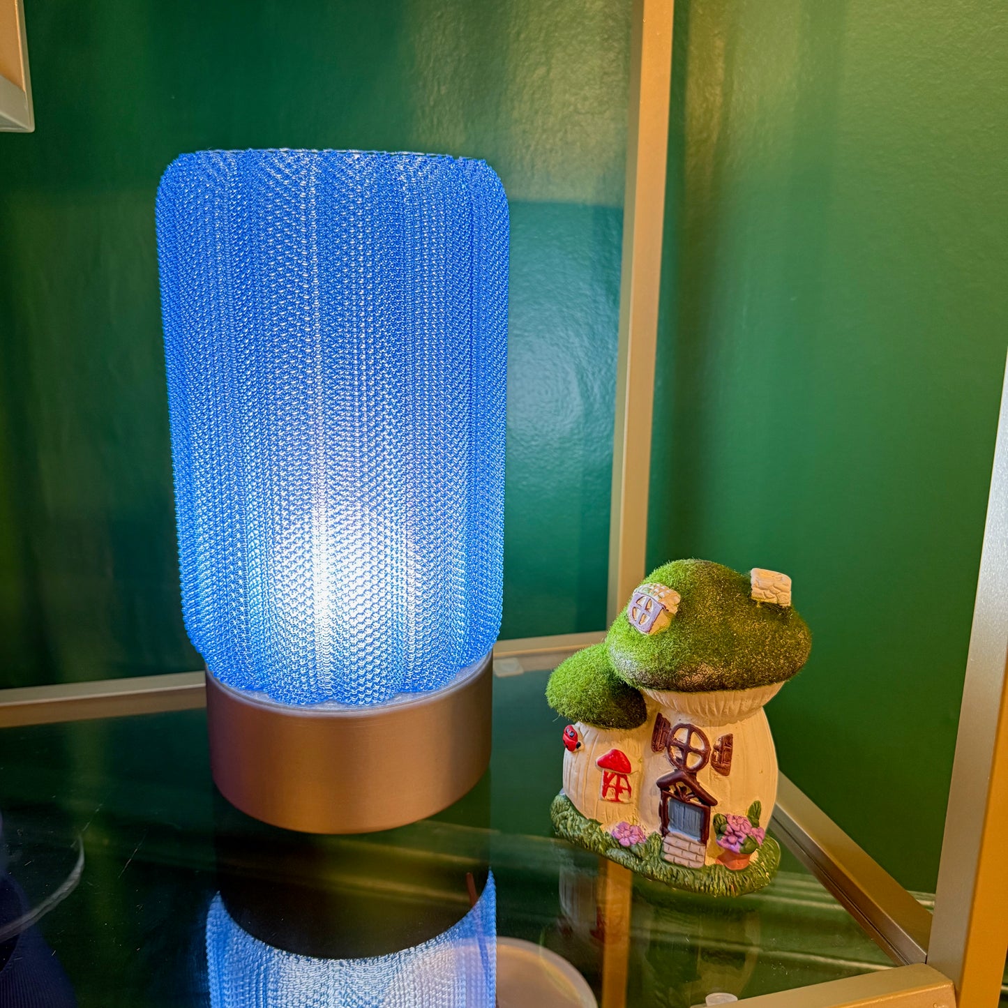 A blue mesh lamp stands beside a small, decorative mushroom house on a glass shelf.