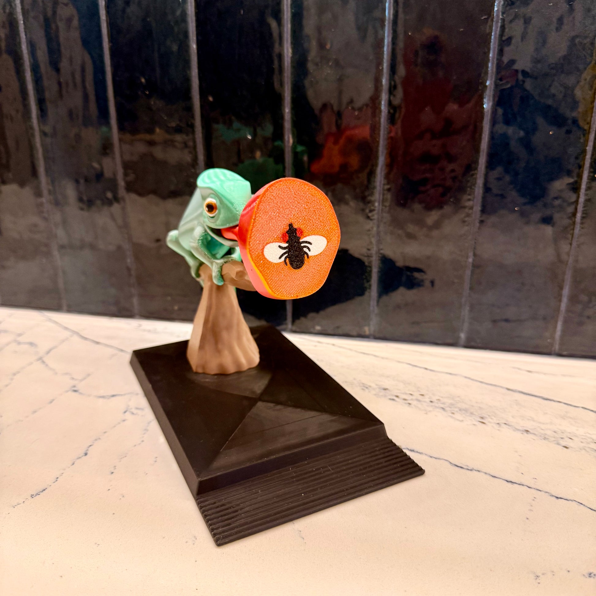 Small figurine of a green creature holding an orange disc with a bee on it, on a reflective surface.