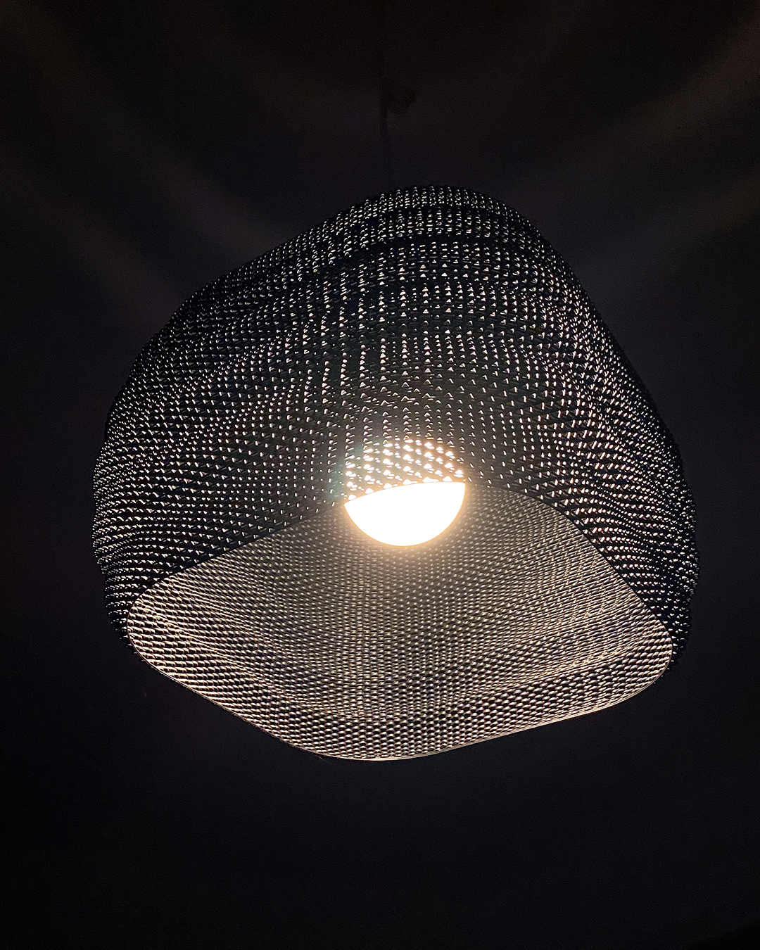 Daira Lampshade beautifully illuminated, surrounded by a dark backdrop, highlighting its intricate woven texture.