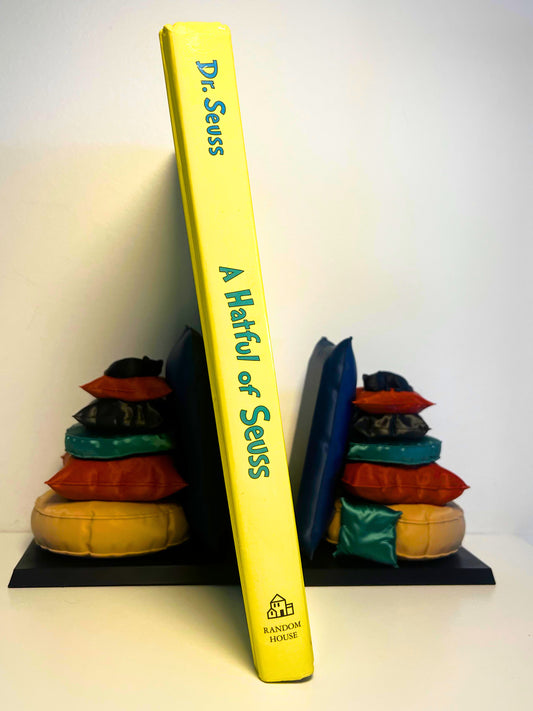 Close-up view of Dr. Seuss's book "A Hatful of Seuss" displayed upright next to colorful, cozy pillow stack bookend, adding whimsy to any bookshelf.
