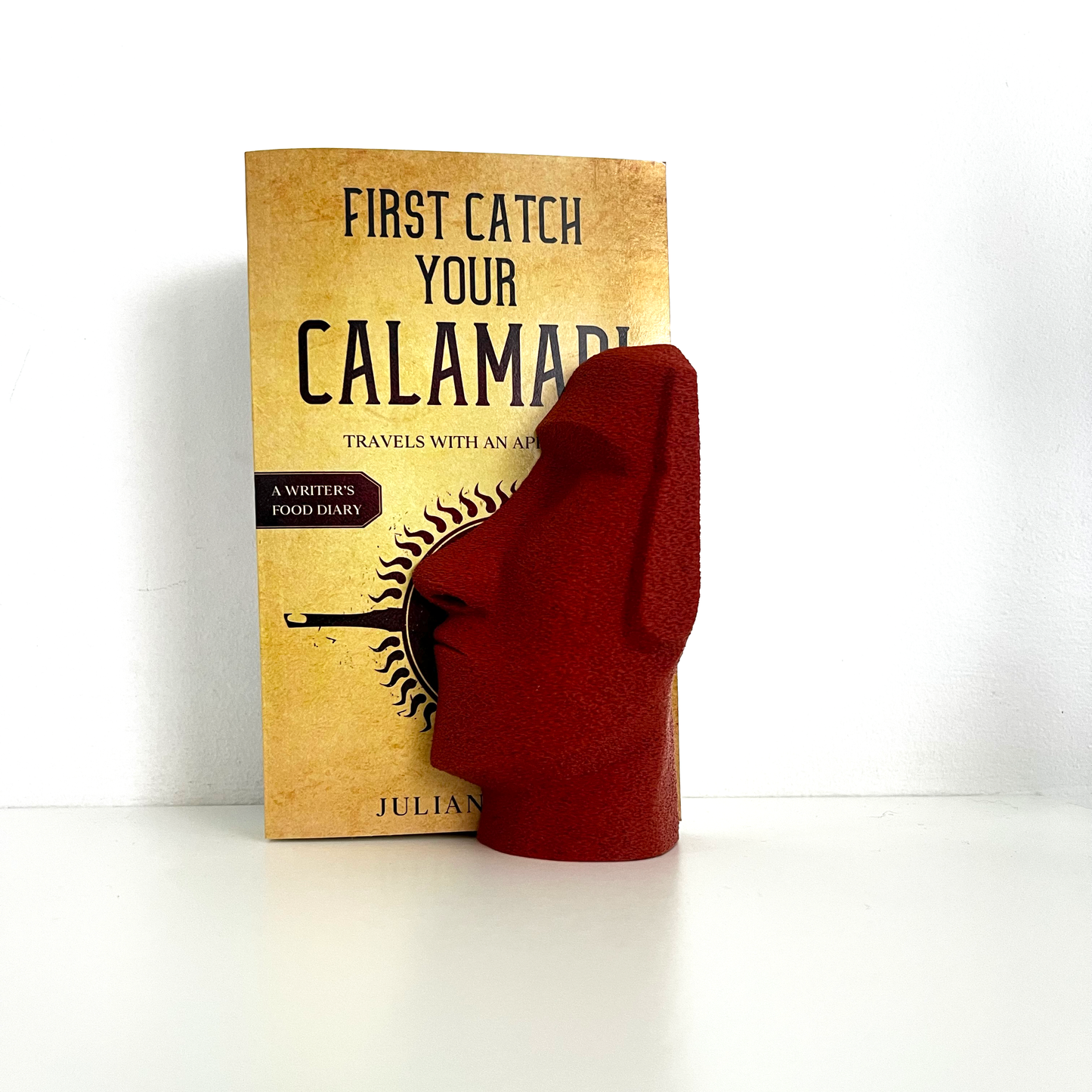 Red Moai Bookend next to the book 'First Catch Your Calamari,' showcasing its unique sculptural features as a stylish bookshelf accessory.
