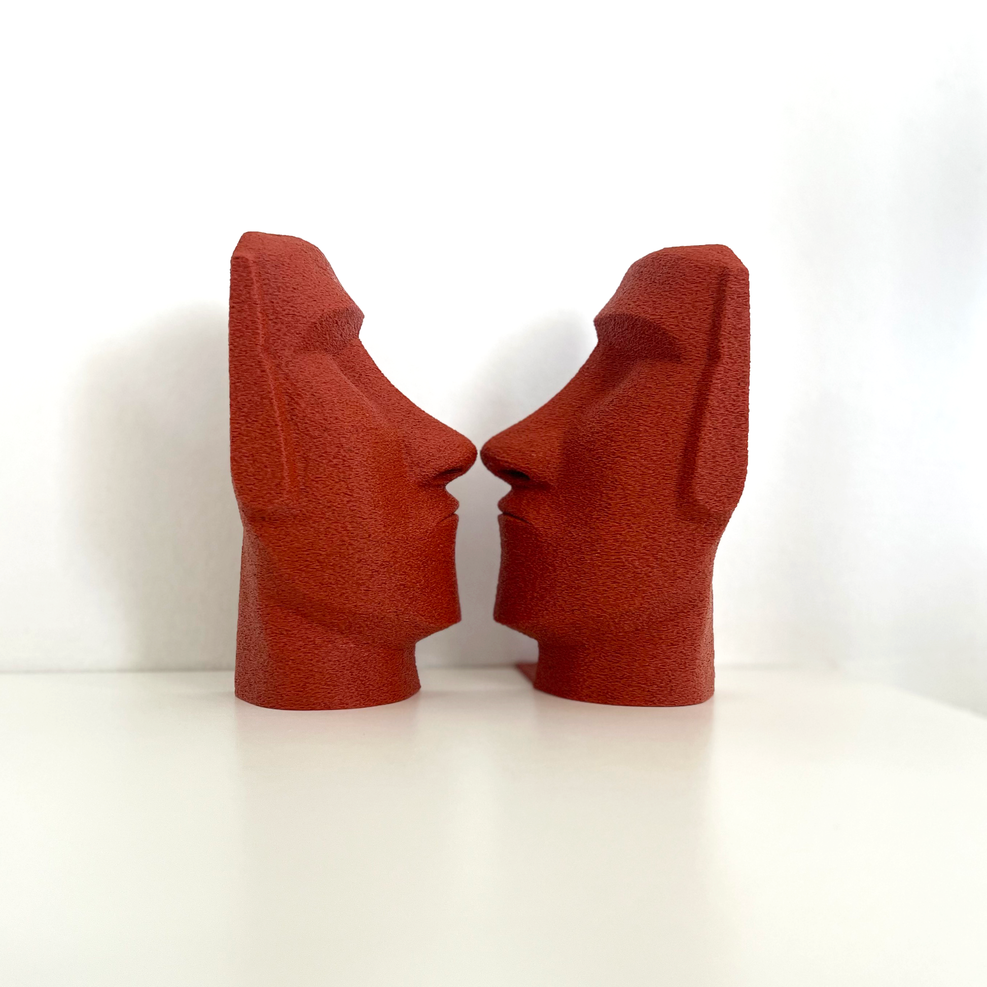 A pair of Moai Bookends in striking red color, positioned to face each other against a plain white backdrop, showcasing their sculptural appeal for bookshelves.