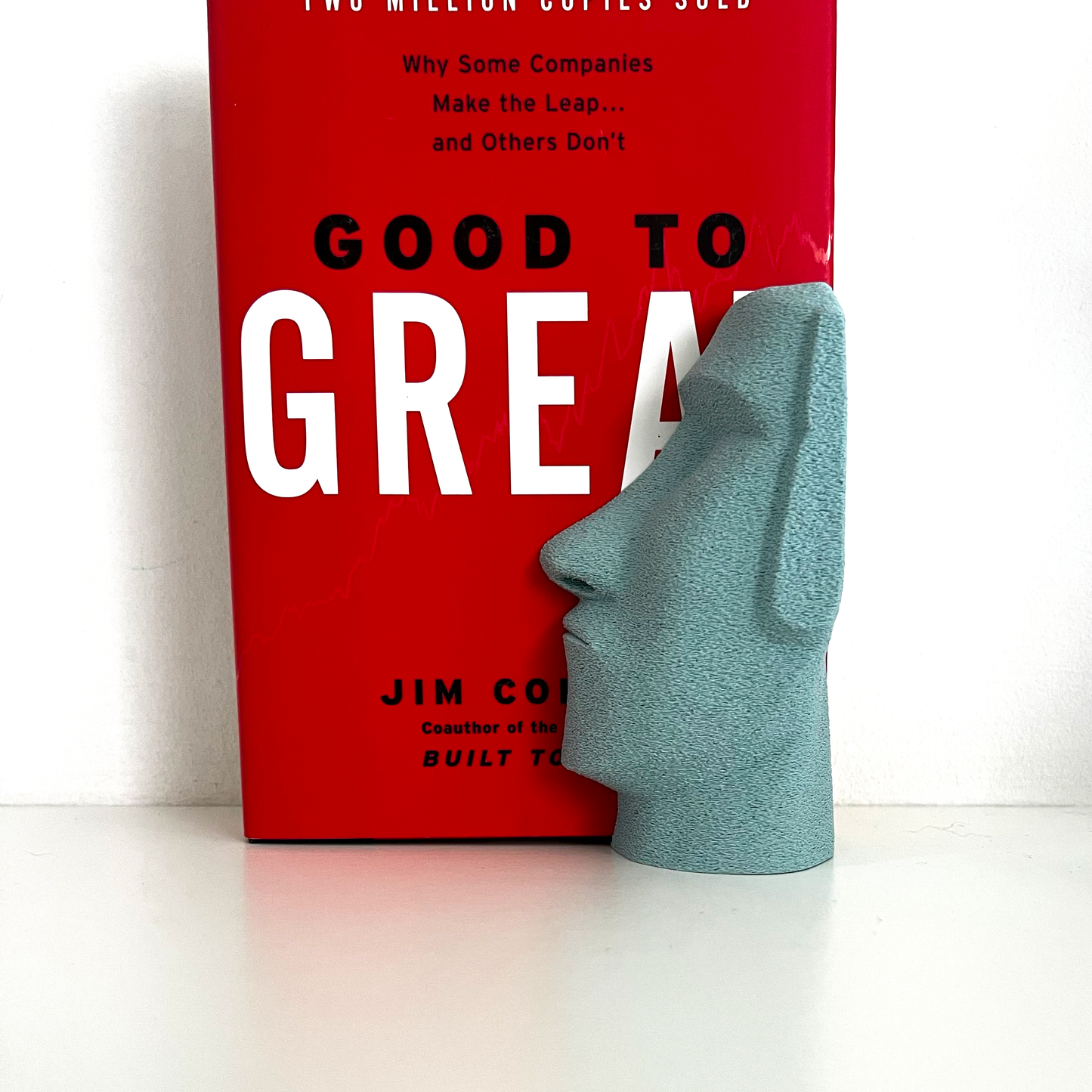 Light blue Moai Bookend placed next to the book 'Good to Great,' highlighting its sculptural design and functionality on a minimalist shelf.
