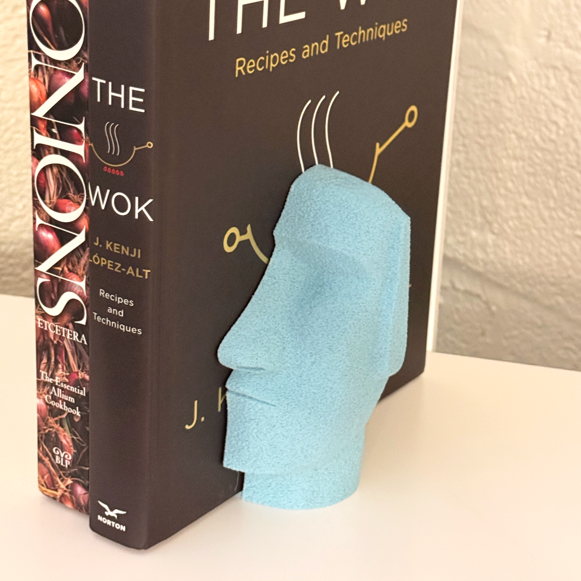 A light blue Moai Bookend accentuating the book 'The Wok' on a cozy shelf, demonstrating the artistic charm and organization it brings.