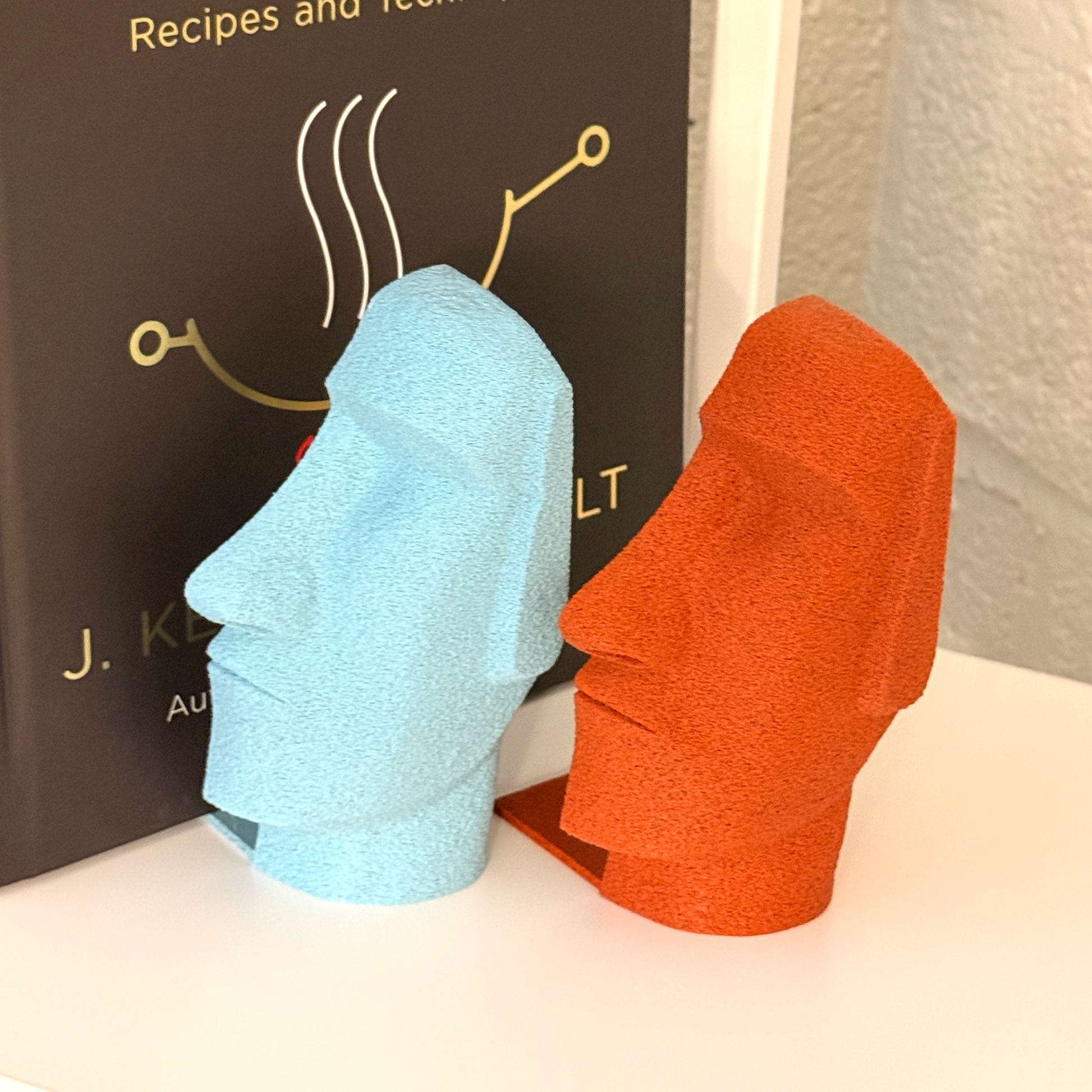 Pair of Moai Bookends in light blue and red, positioned near 'The Wok,' illustrating their distinctive designs while enhancing the bookshelf aesthetic.