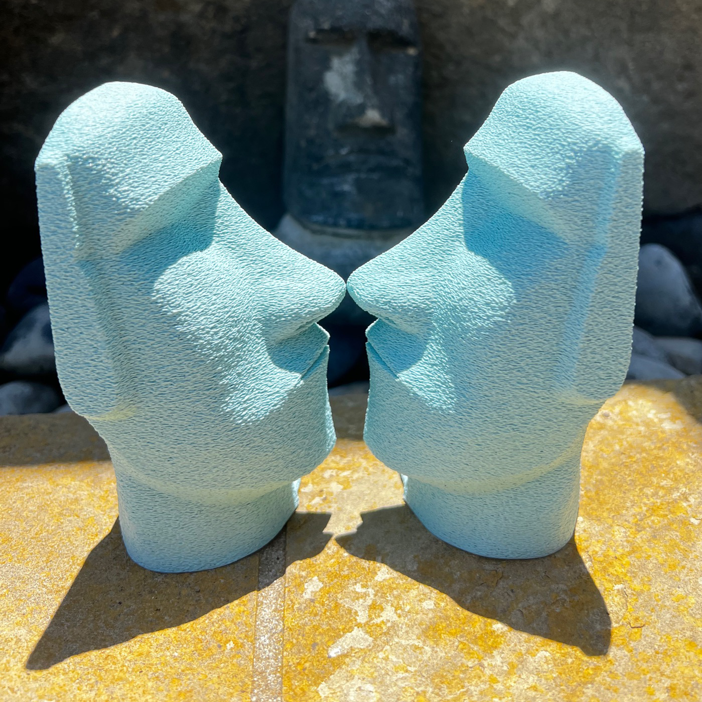 Two Moai Bookends in a vibrant light blue color, facing each other on a stone surface, emphasizing the unique design that enhances any bookshelf.