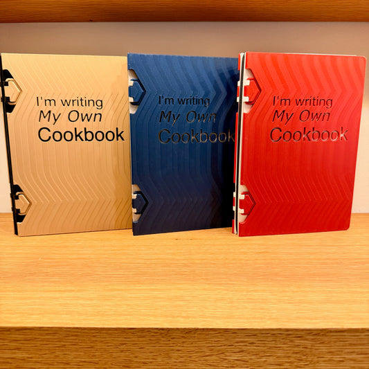 Three stylishly designed 'I Am Writing My Own Cookbook' custom recipe books in beige, blue, and red on a wooden shelf, perfect for organizing cherished family recipes.