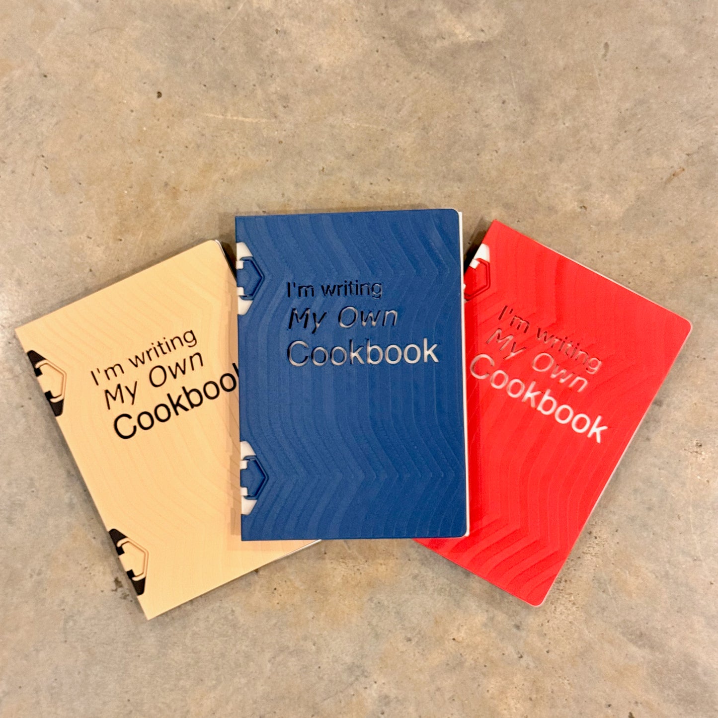 Close-up of three 'I Am Writing My Own Cookbook' custom recipe books in beige, blue, and red, showcasing their attractive cover designs ideal for food enthusiasts.